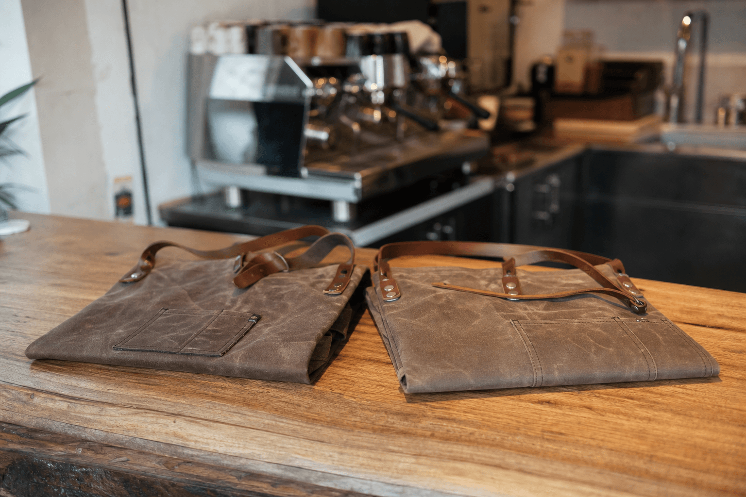Waxed canvas barista half aprons with leather straps on a cafe counter