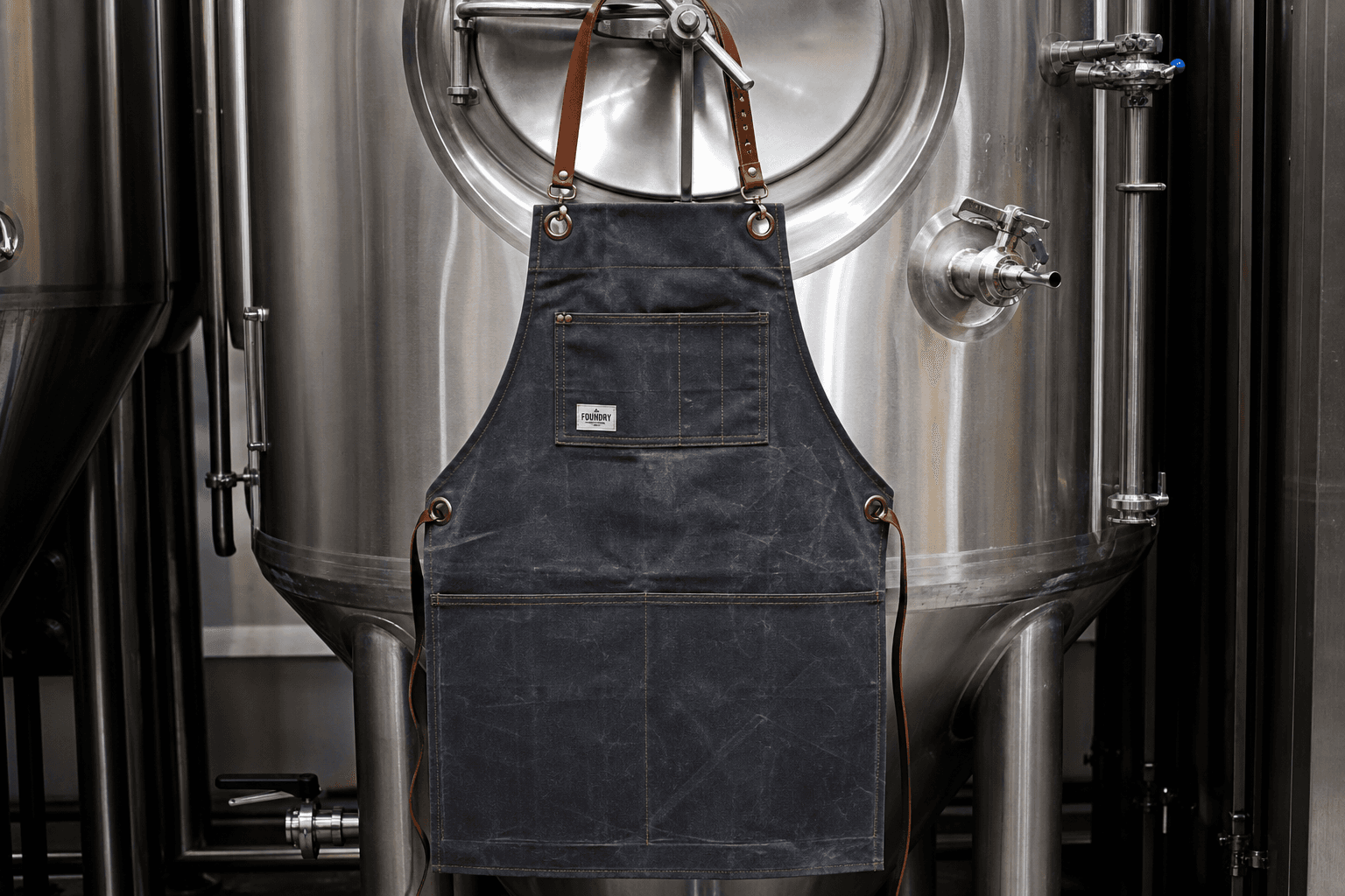 A waxed-canvas brewery apron with leather straps on a steel tank backdrop