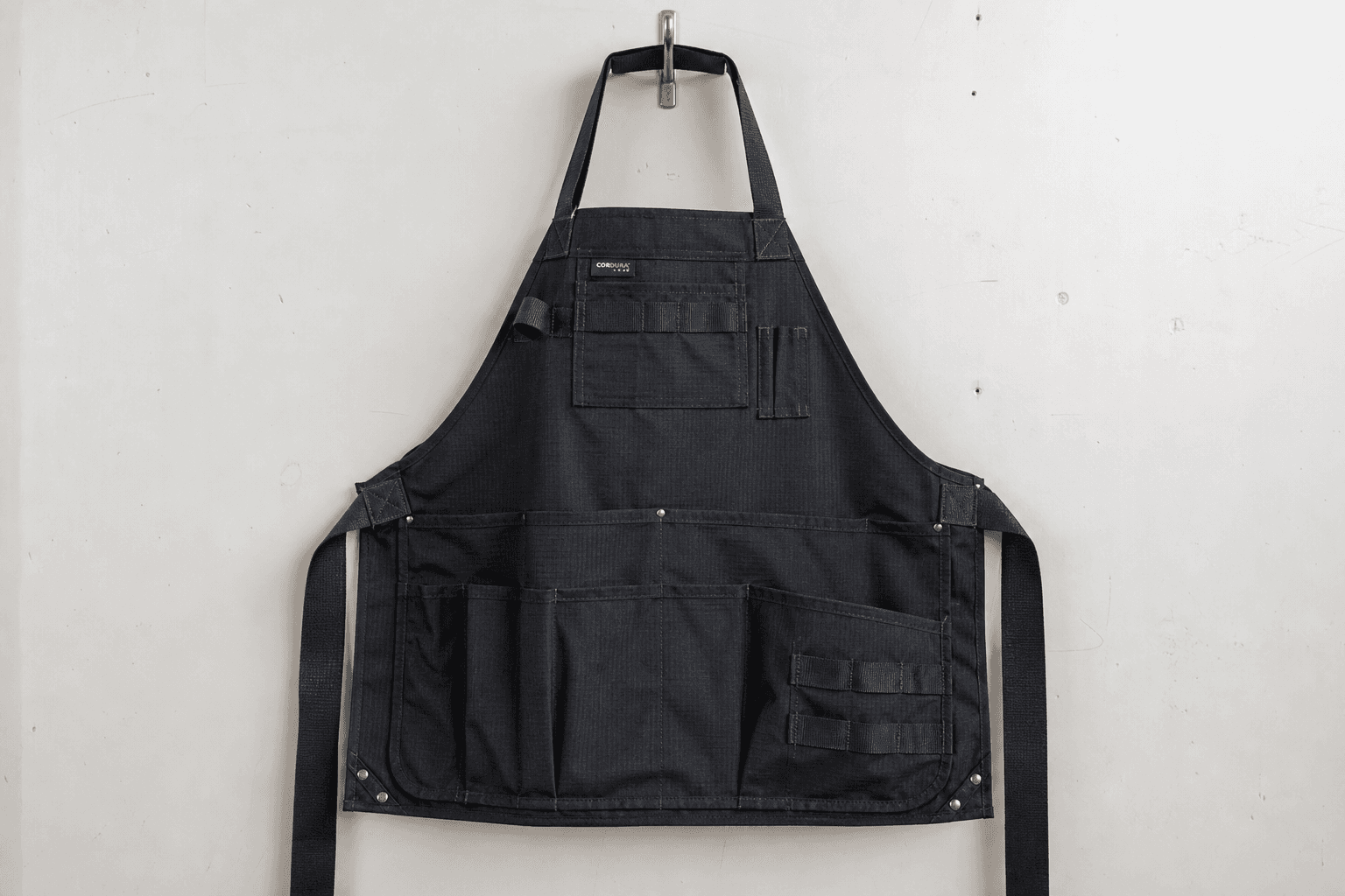 A black Cordura ripstop work apron with reinforced pockets on a metal hook
