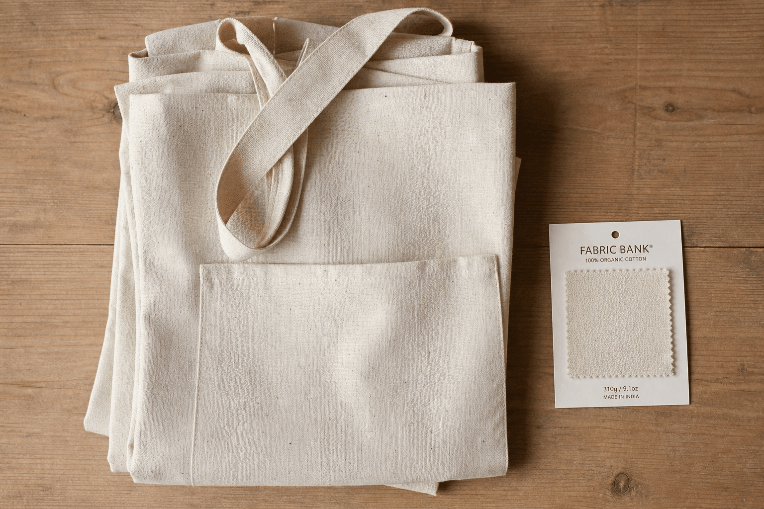 Folded undyed organic cotton aprons with a GOTS swatch card on a wooden table